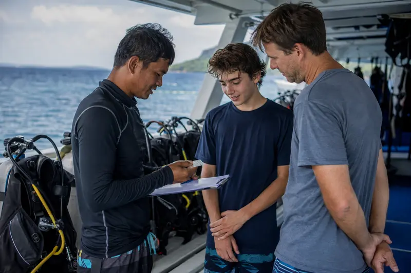 A liveaboard crew member checking a teenage diver's certification with a guardian present